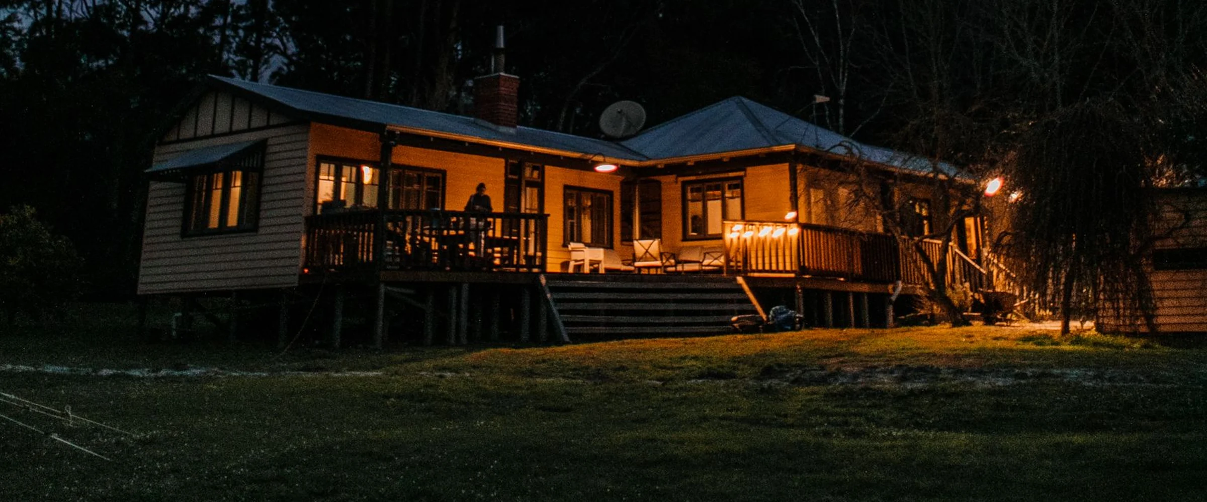 A wood-sided house with its porch lights on at dusk, tall trees around it